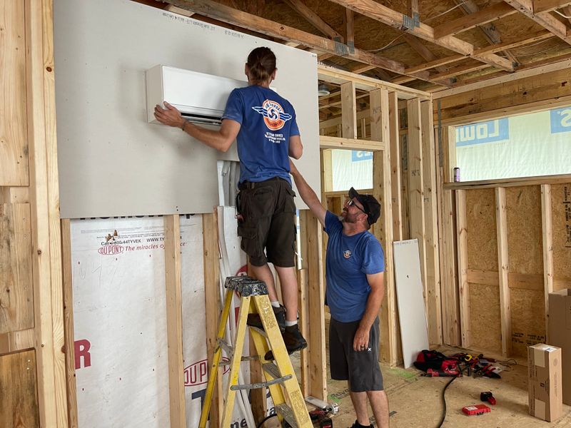 Two Air Support technicians installing a ductless mini-split indoor unit on a ladder in new construction