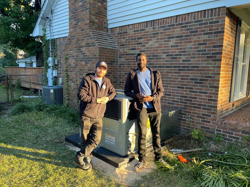 Two Air Support technicians standing next to a completed condenser installation at a brick home